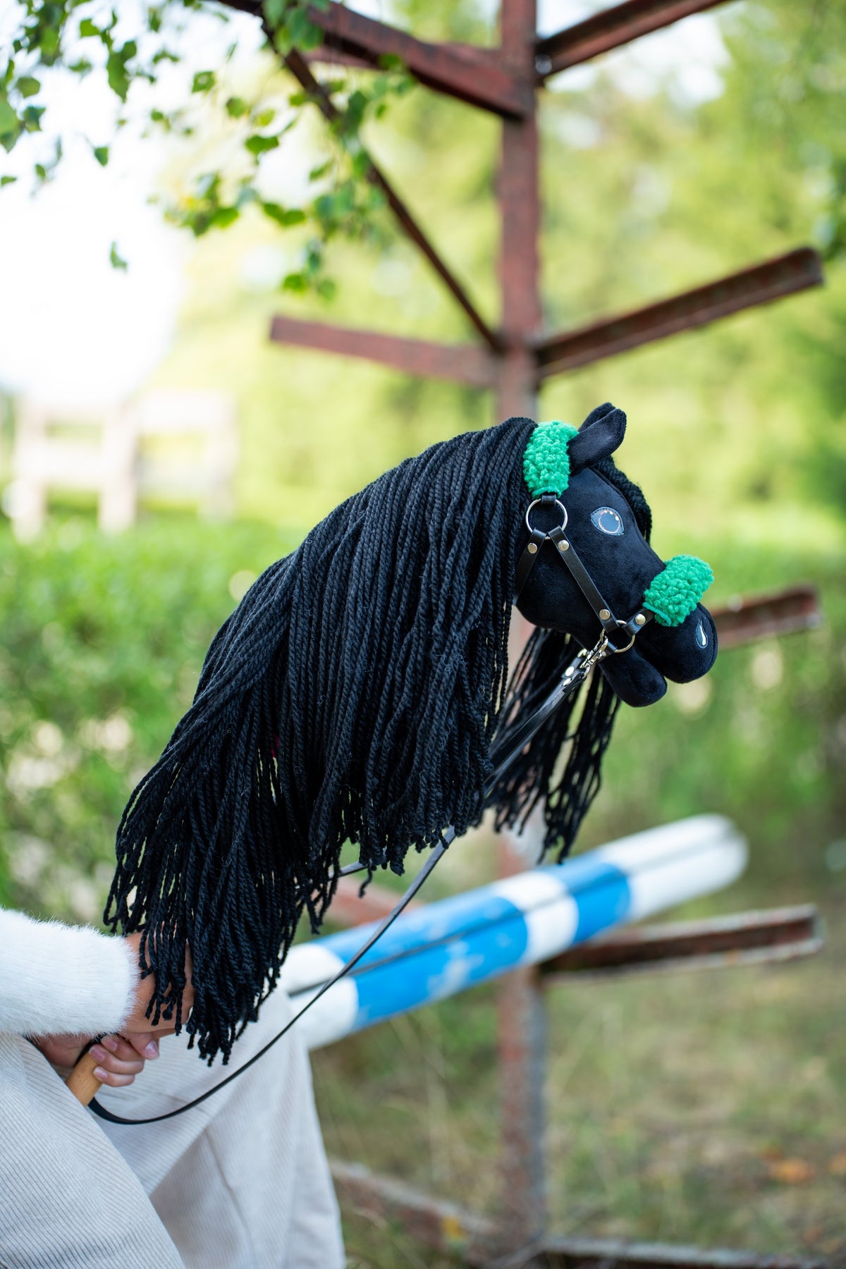 Black Hobby Horse with a long yarn mane, green fur halter and black reins by a blue and white jump pole