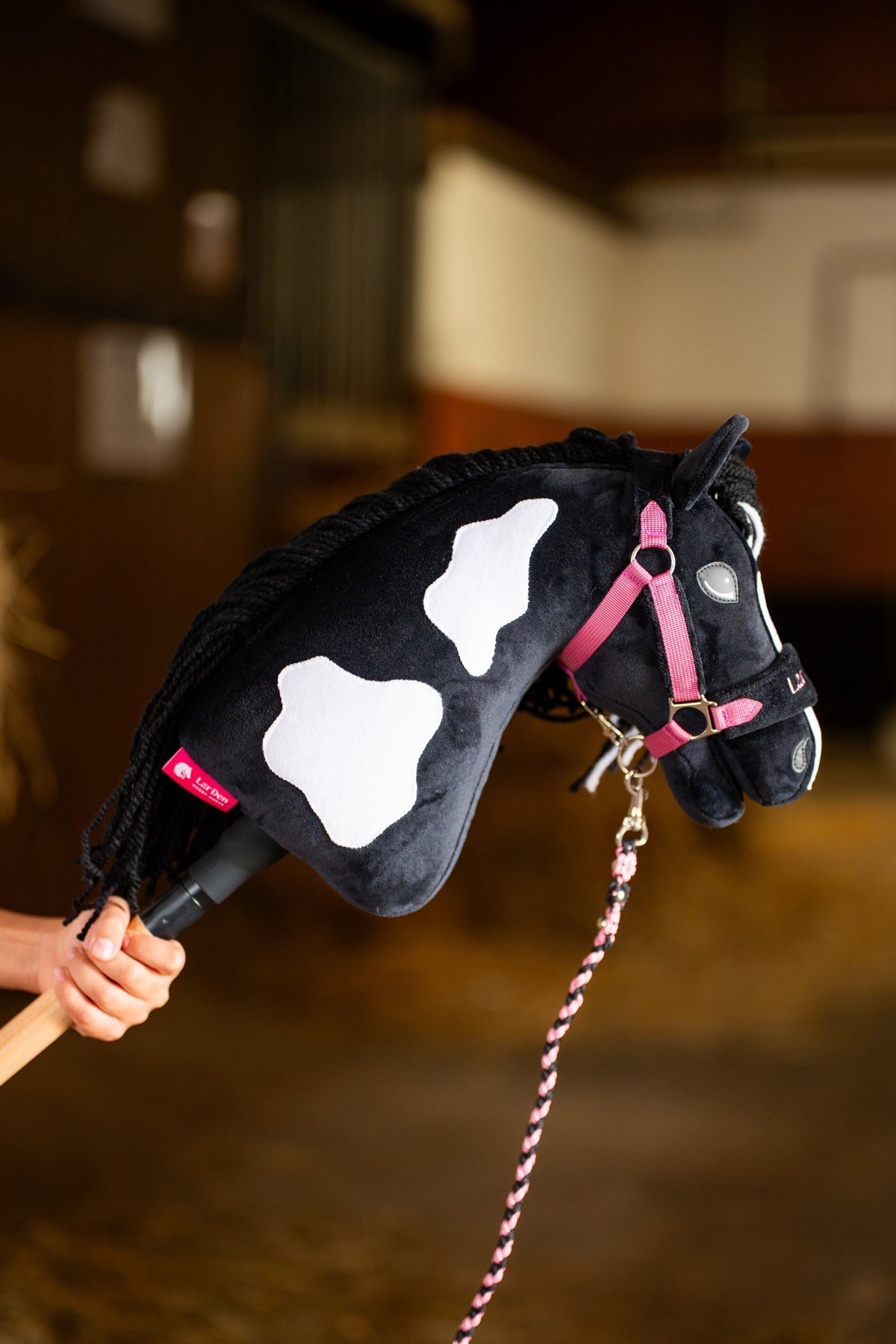 Nagy fehér foltos Hobby Horse Merlin rózsaszín hevederes kötőfékben, rózsaszín-fekete fonott vezetőszárral egy istállóban