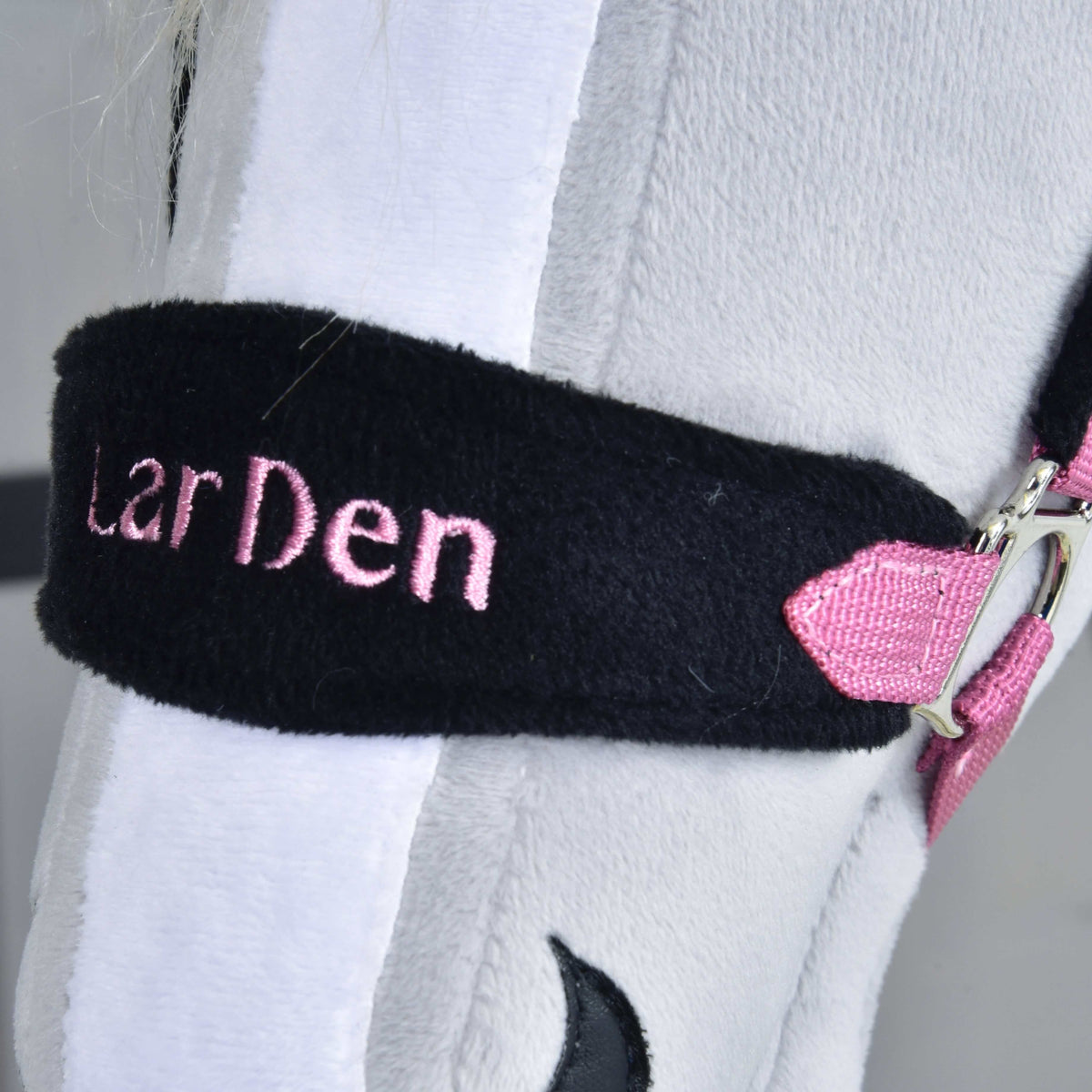 A fekete szőrmés orrszíj közelről rózsaszínnel hímzett LarDen felirattal egy fehér hókás, világosszürke Hobby Horse lovon