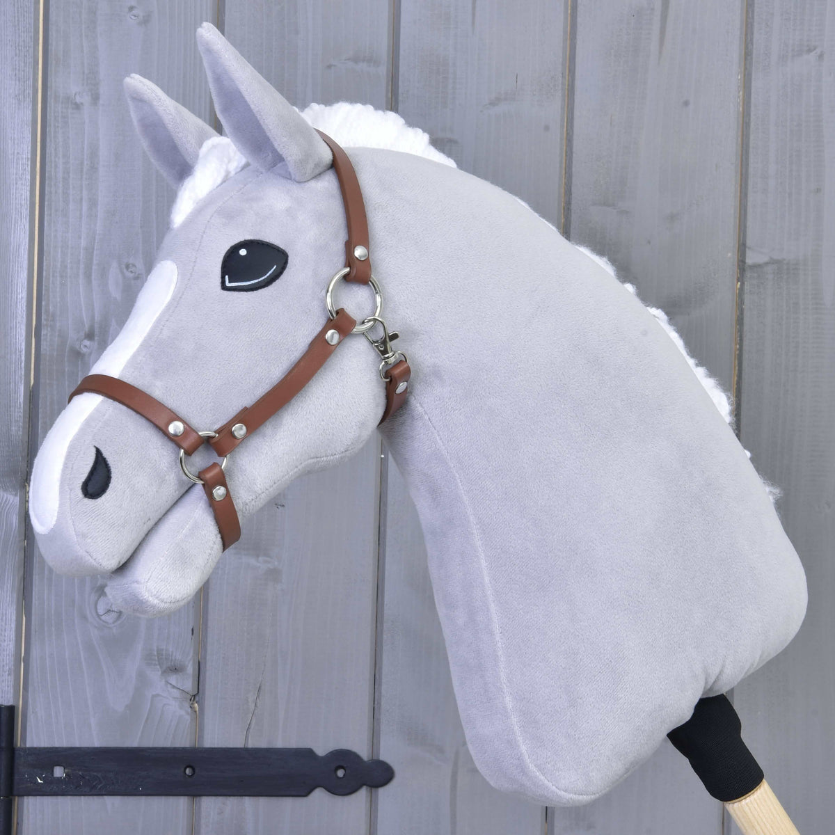 Grey plush Hobby Horse with a white mane and blaze wearing the brown leatherette halter in front of a grey wooden wall