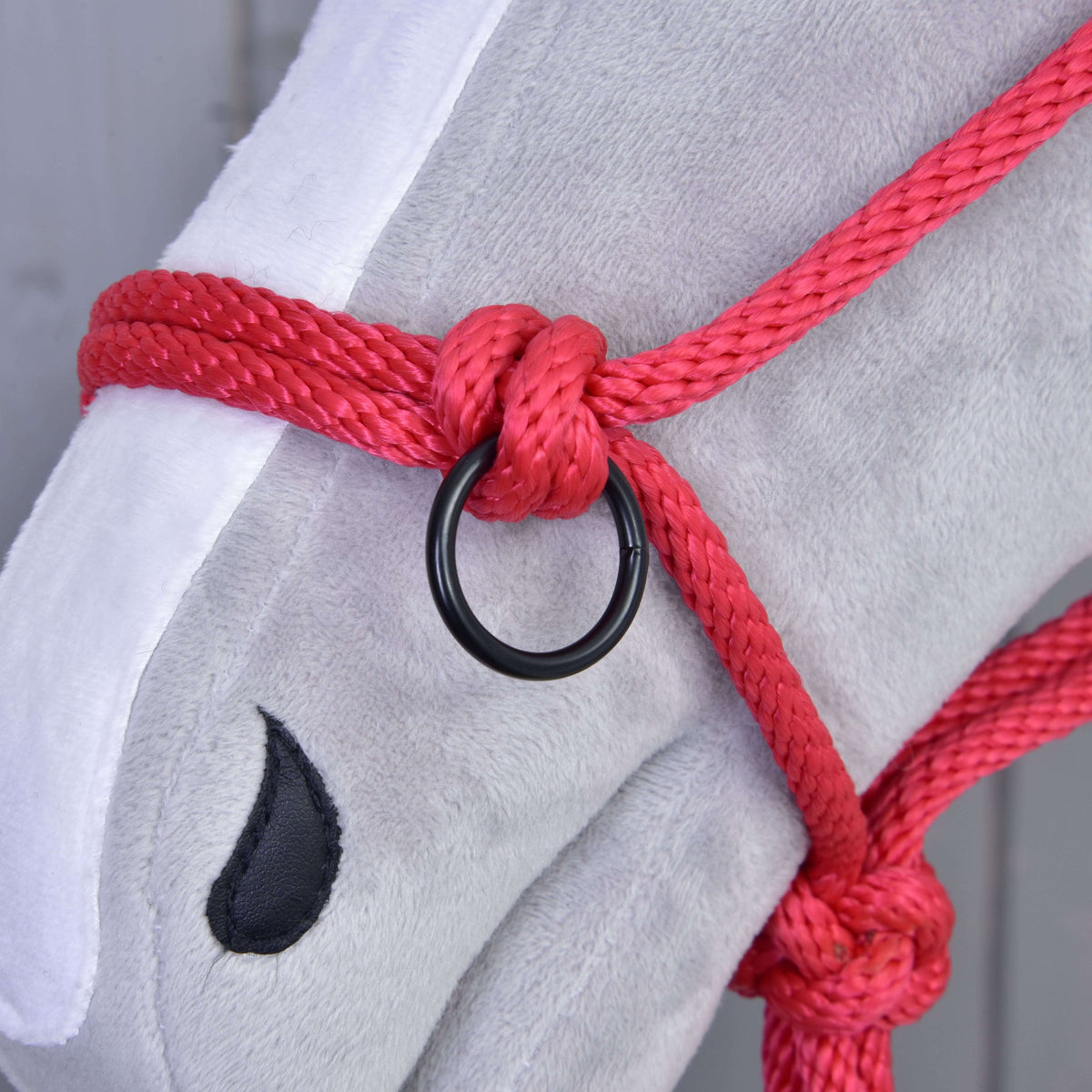 A piros csomózott kötőfék egyik csomója és fekete karikája közelről egy szürke Hobby Horse ló fehér hókáján