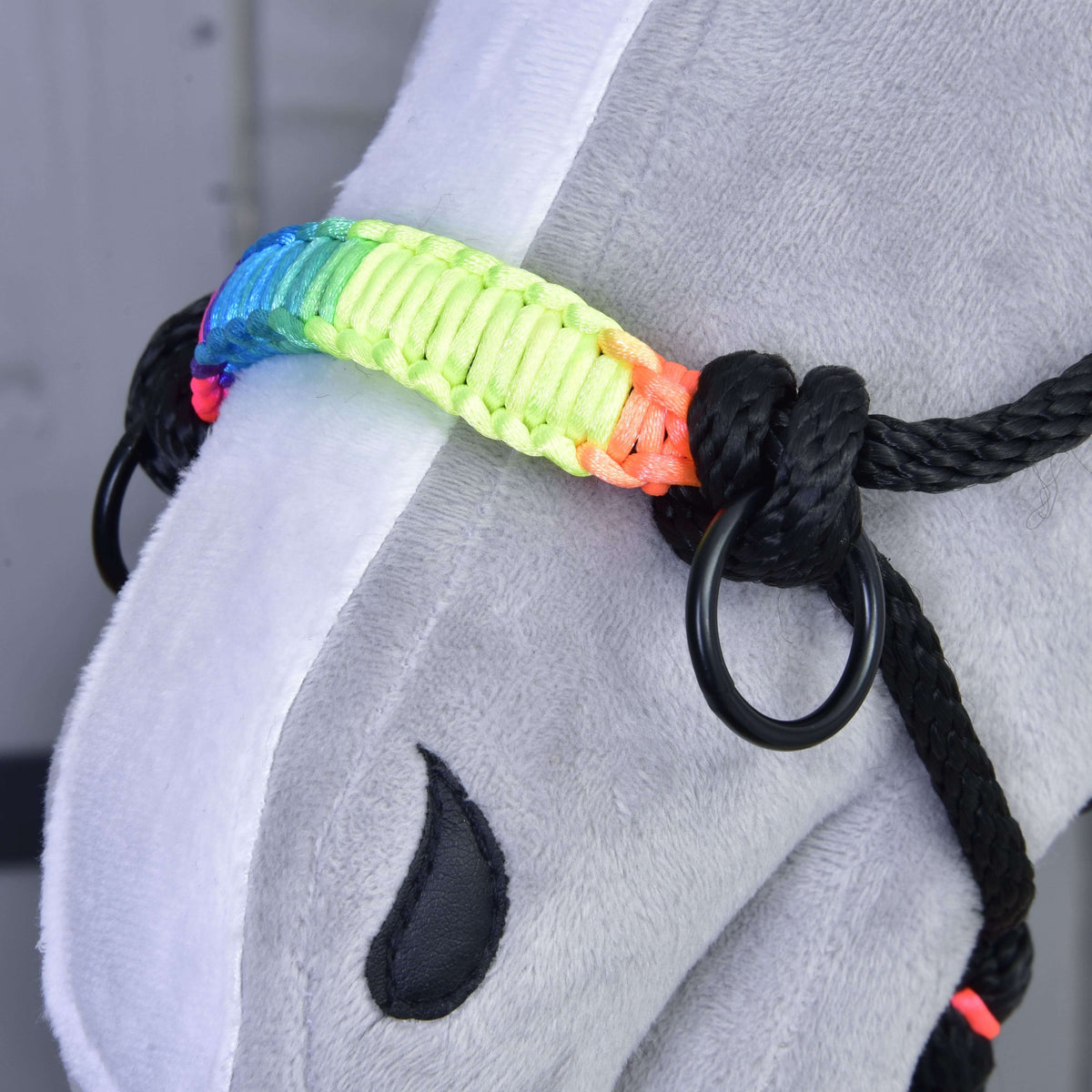 Közeli kép a csomózott kötőfék szivárványos fonott orrszíjáról, fekete karikájáról és csomóiról egy szürke plüss Hobby Horse lovon