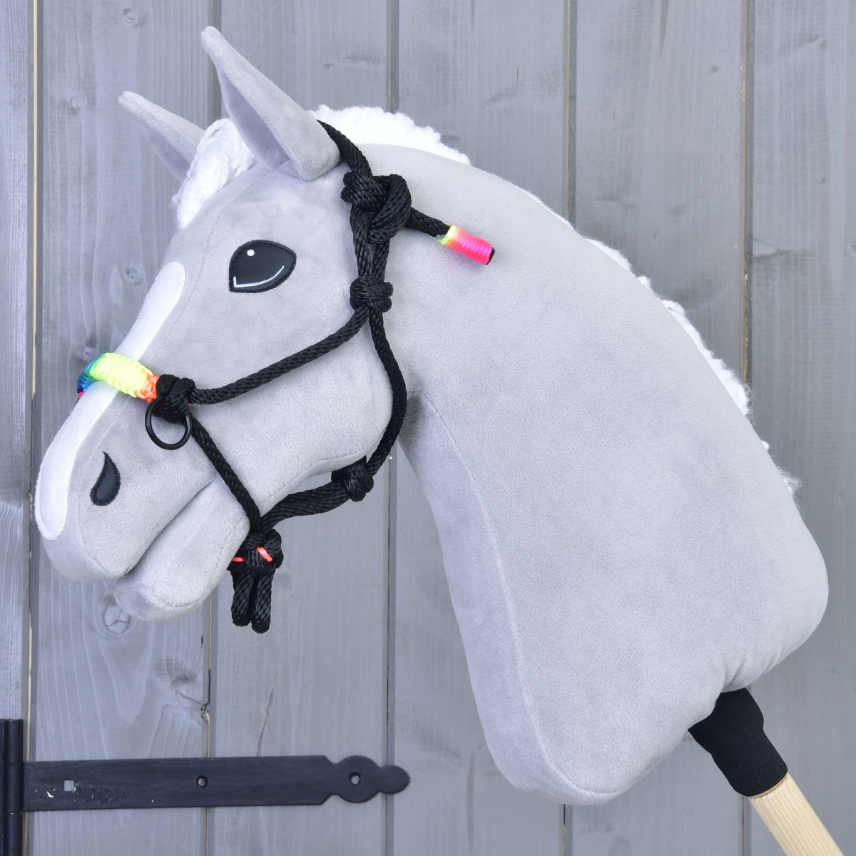 Szürke plüss Hobby Horse ló fehér sörénnyel, a fekete csomózott kötőfékben szivárványos orrszíjjal, szürke fafal előtt