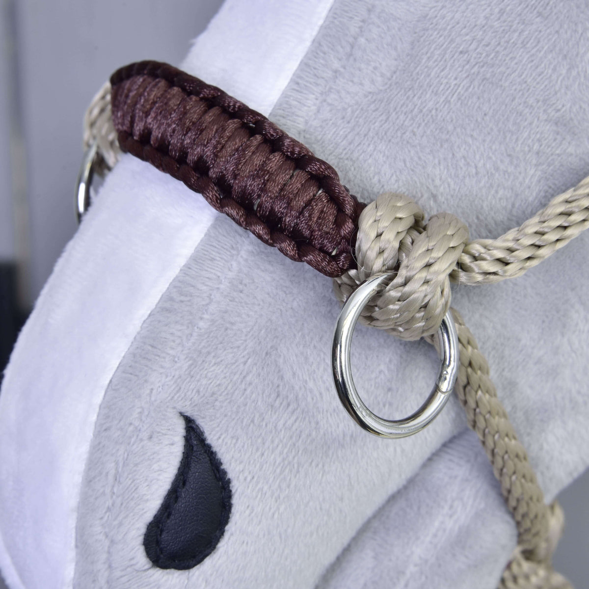 Közeli kép a bézs csomózott kötőfékről barna fonott orrszíjjal és ezüstszínű karikával egy szürke plüss Hobby Horse lovon