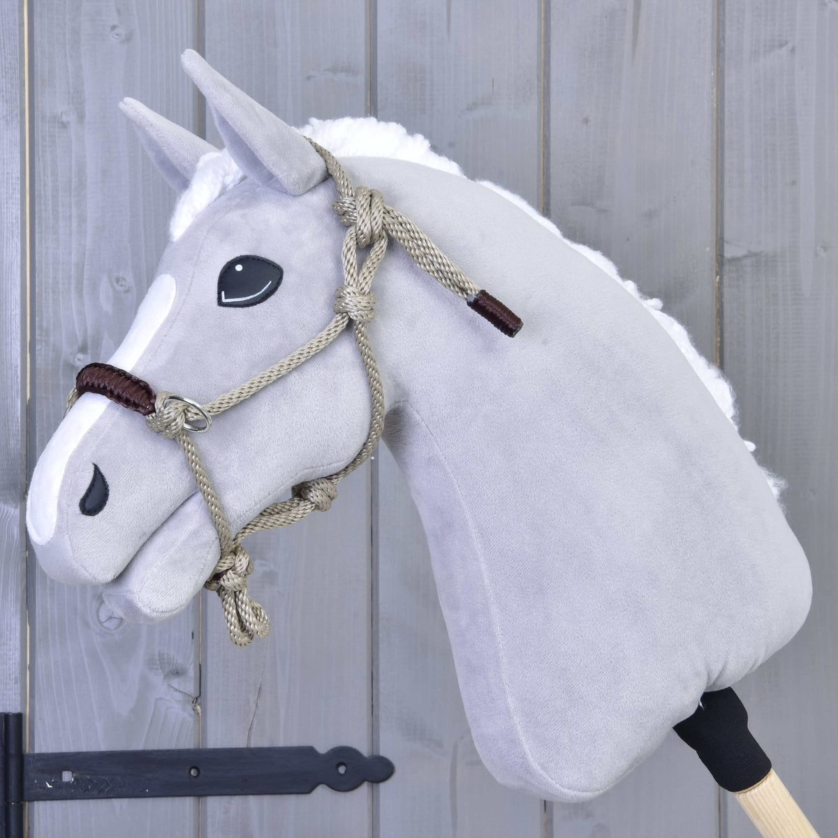 Szürke plüss Hobby Horse ló fehér sörénnyel, a bézs csomózott kötőfékben barna orrszíjjal, szürke fafal előtt