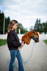 Sötét pulóveres kislány homokos úton vezeti a Hobby Horse Pippa lovat kékeszöld kötőfékben és kék vezetőszáron