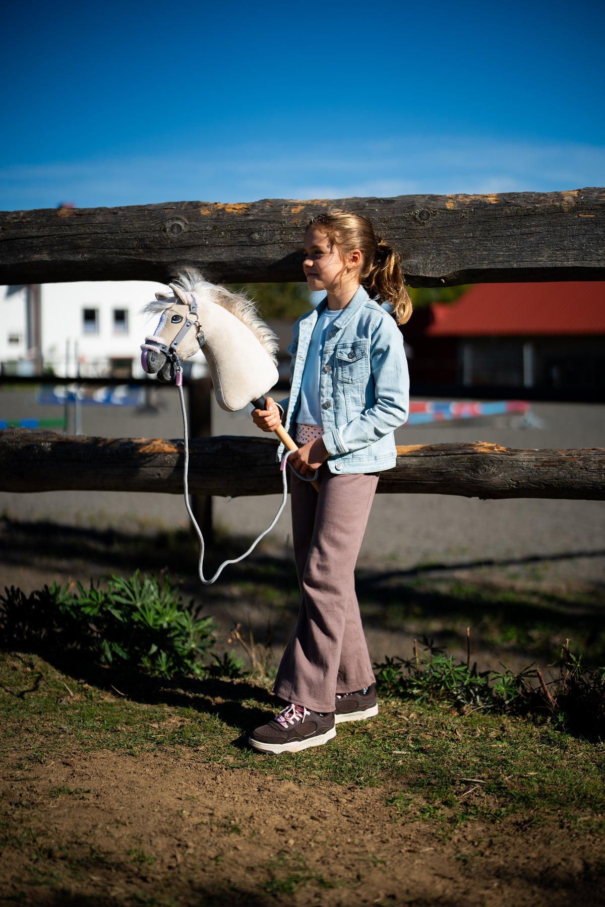 Farmerdzsekis kislány egy rönkkerítésnél, kezében a Hobby Horse Pixie ló szürke szőrmés kötőfékben, vezetőszárral