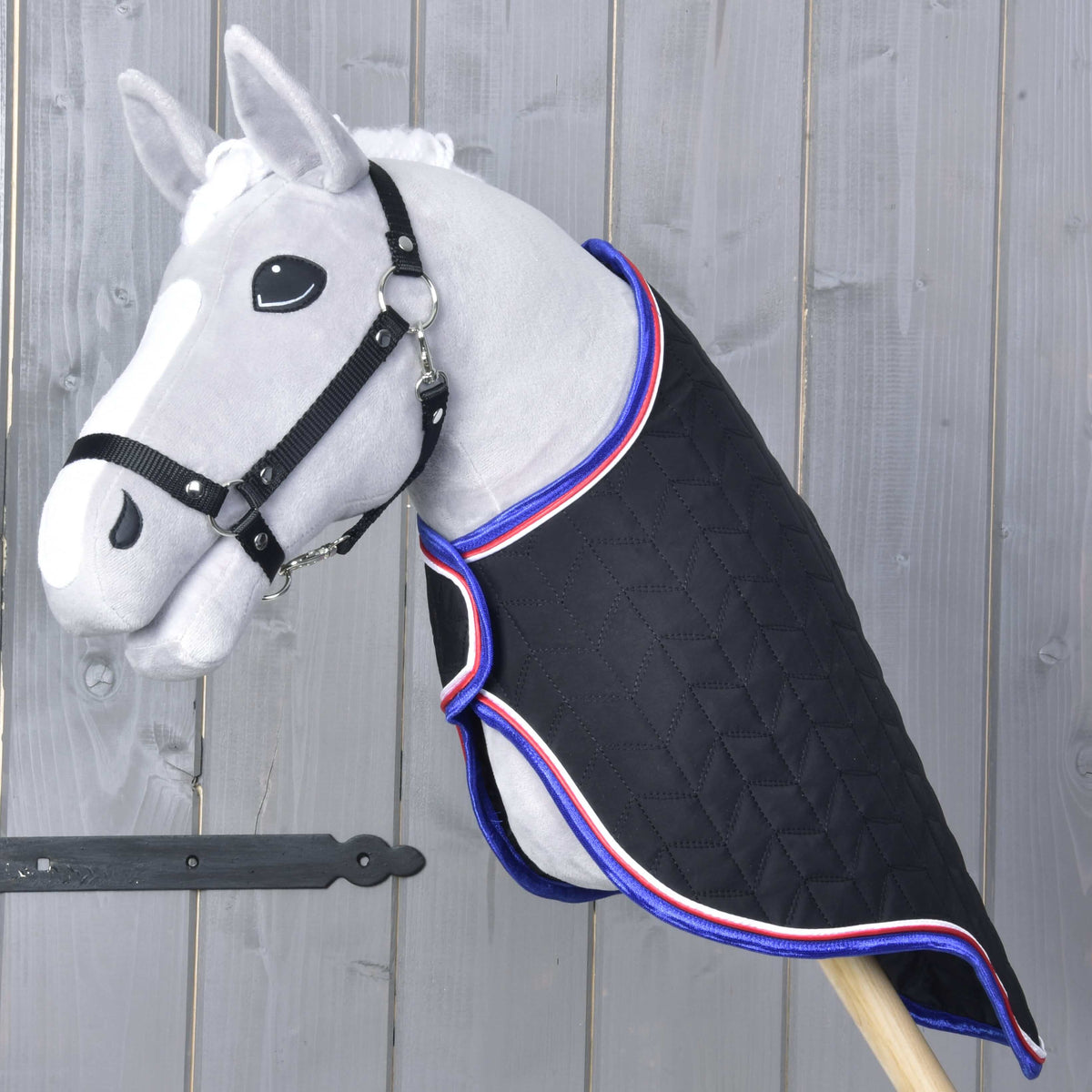 Szürke Hobby Horse ló fekete kötőfékben, fekete steppelt szállítótakaróban kék, piros és fehér szegéllyel, háromnegyedes nézetből