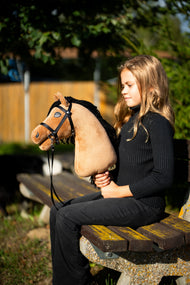 Fekete pulóveres lány ül egy parkpadon az esti napsütésben, és a Hobby Horse Samantha lovat tartja fekete kantárban, szárral