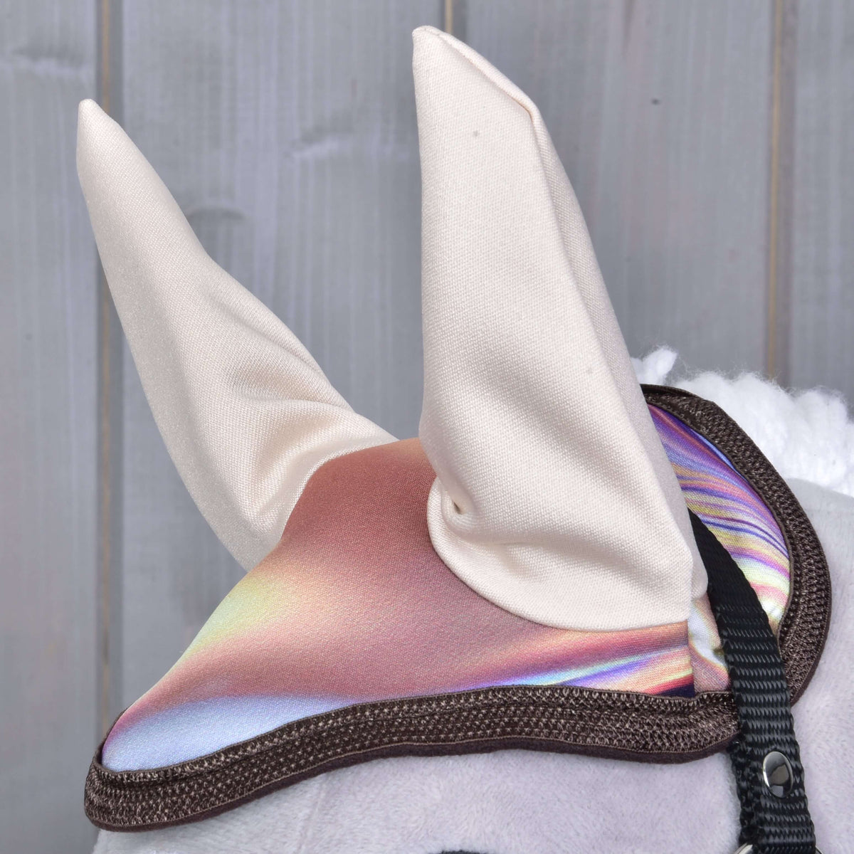 A fülvédő közelről magas krémszínű fülekkel és csillámló rózsaszín-orgonalila, barna szegélyű fejrésszel egy szürke Hobby Horse lovon
