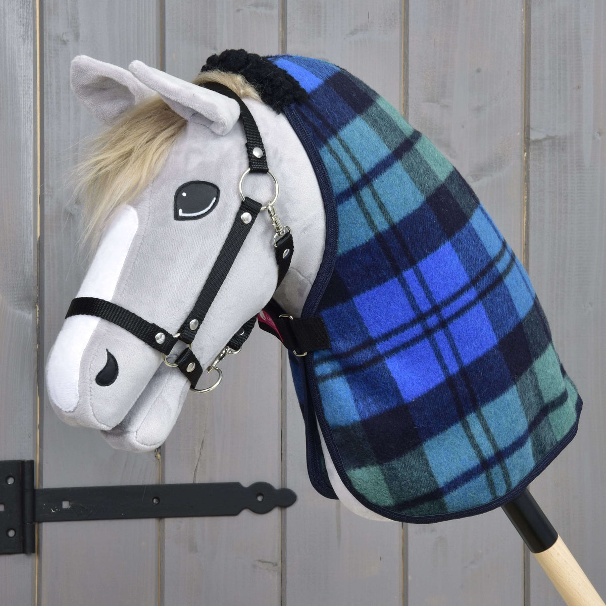 Szürke plüss Hobby Horse ló fekete kötőfékben, a kék-zöld skótkockás istállótakaróban, szürke fafal előtt