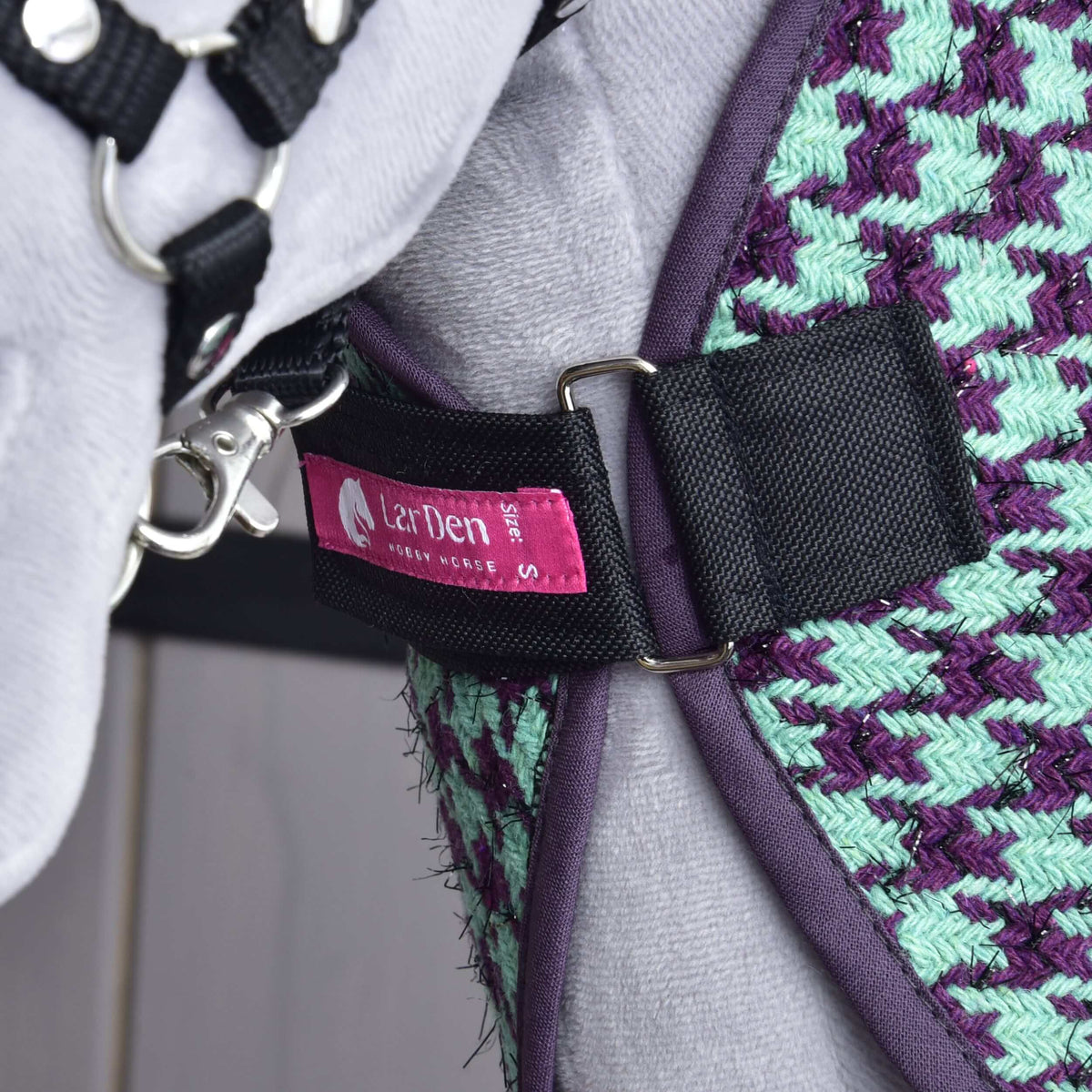 A zöld Hobby Horse istállótakaró mellpántja D-karikákkal bekapcsolva egy szürke plüss mellkason, látszik a rózsaszín LarDen címke