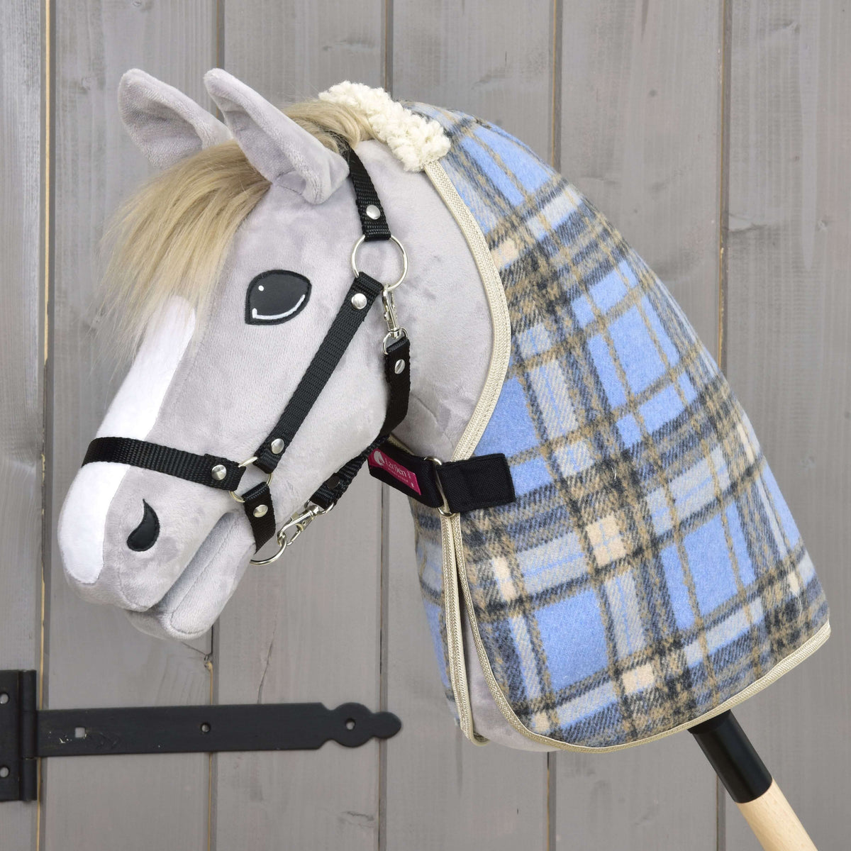 Szürke plüss Hobby Horse ló fekete kötőfékben, a világoskék kockás istállótakaróban, bezárt fekete mellszíjjal