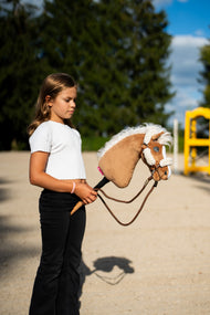 Fehér felsős lány egy homokos pályán a Hobby Horse Star lovat tartja, itt krémszínű szőrmés barna kötőfékben és barna kötél kantárszárral