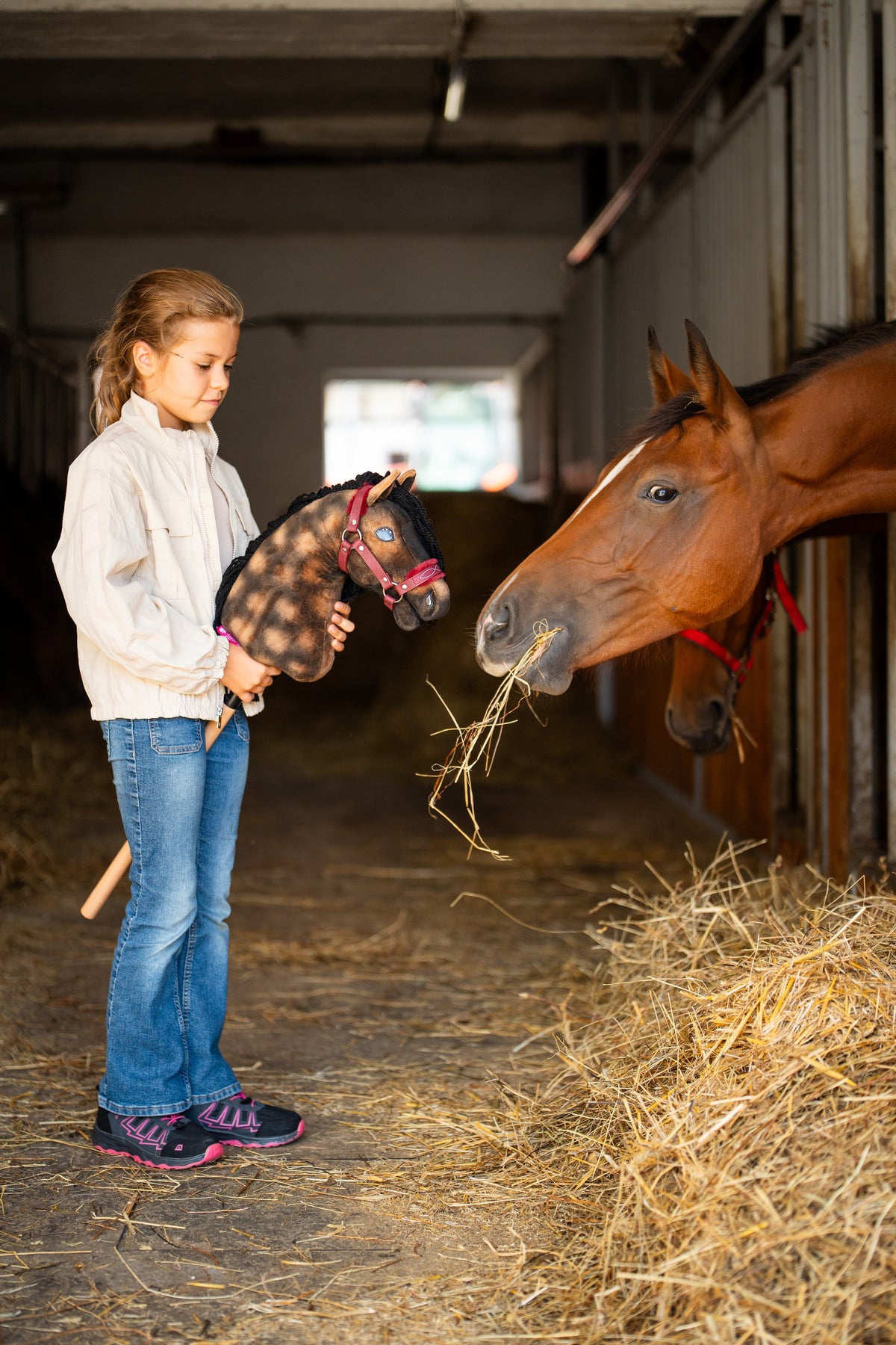 Lány az istállóban egy almás barna Hobby Horse lovat tart bordó szőrmés kötőfékben, mellette egy szénát evő pej ló