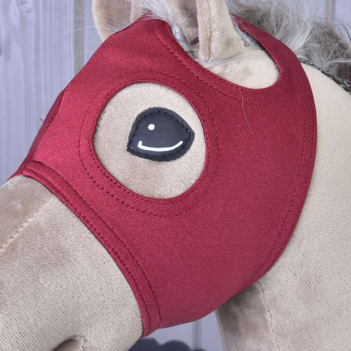 Close-up of the burgundy mask without ears on a beige Hobby Horse, showing the stitched round eye opening and ear hole