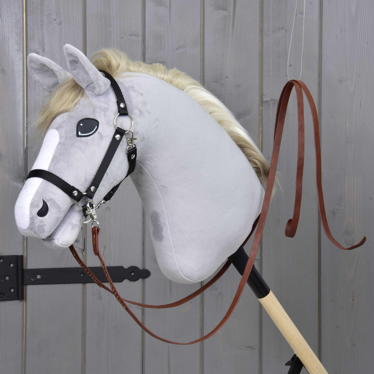 Világosszürke Hobby Horse ló fekete kötőfékkel, amelyre a Dark Brown osztott western kantárszár van csatolva, egy szürke faajtó mellett