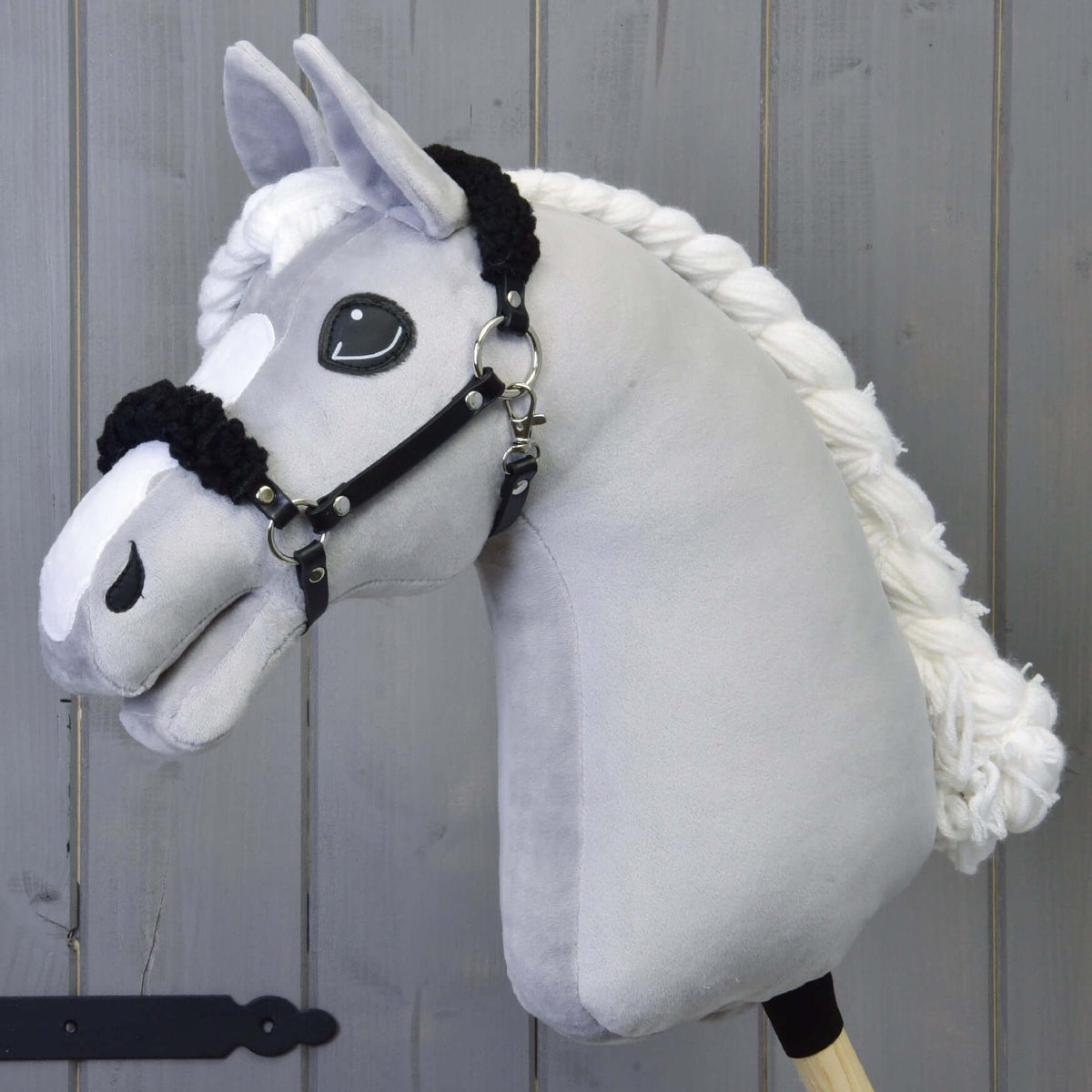 Grey plush Hobby Horse with a braided white mane wearing the black halter with black fur in front of a grey wooden wall