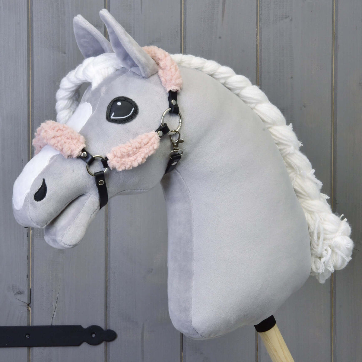 Grey plush Hobby Horse with a braided white mane wearing the black halter with old rose fur in front of a grey wooden wall
