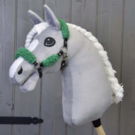 Szürke plüss Hobby Horse ló fonott fehér sörénnyel, rajta a fekete kötőfék zöld szőrmével, szürke fafal előtt