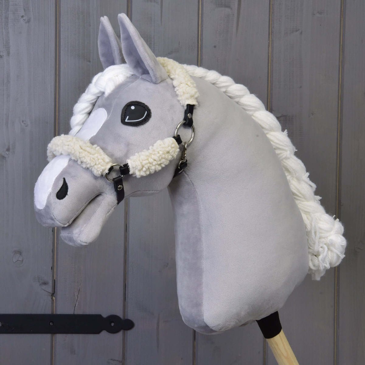 Szürke plüss Hobby Horse ló fonott fehér sörénnyel, rajta a fekete kötőfék teljes krémszínű szőrmeborítással, háttérben fafal