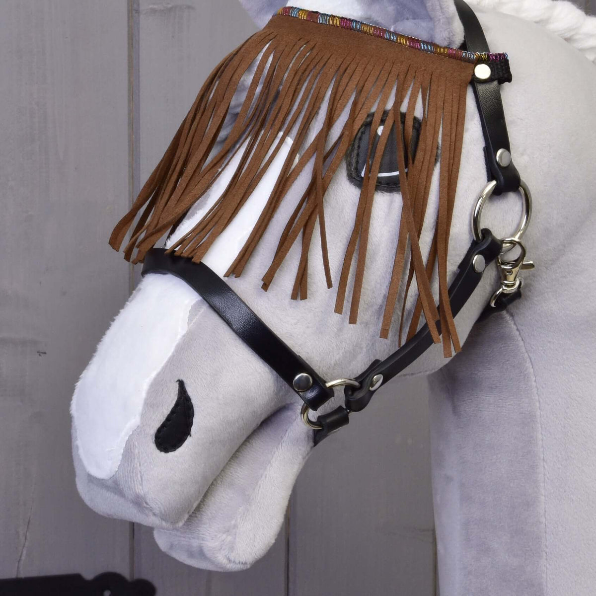 Közeli kép a barna művelúr rojtokról és a színes szövött szegélyről egy szürke Hobby Horse ló fekete kötőfékén