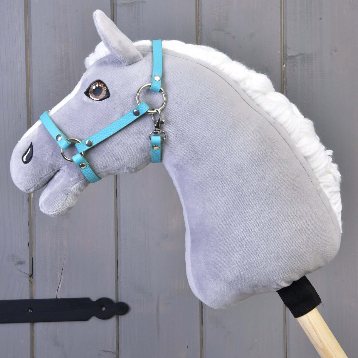 Szürke plüss Hobby Horse ló fehér sörénnyel és barna szemekkel, a türkiz műbőr kötőfékben, szürke fafal előtt