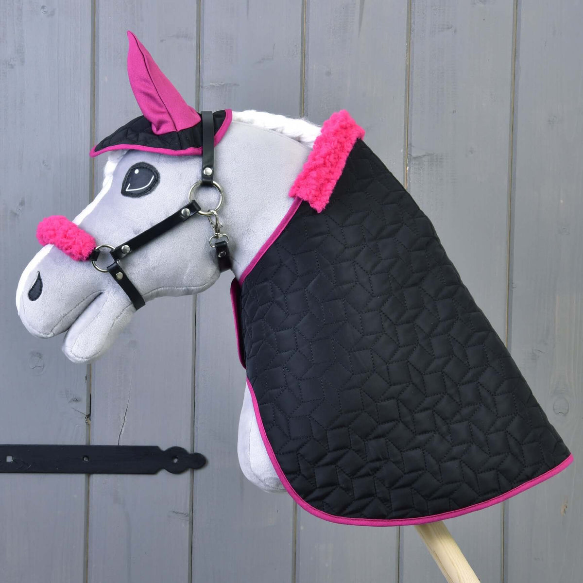 Grey plush Hobby Horse wearing the black quilted blanket, ear bonnet and halter with fuchsia fur, wooden wall