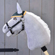 Szürke plüss Hobby Horse ló fekete kötőfékkel, rajta a zöld steppelt fülvédő mustársárga szegéllyel, háttérben fafal